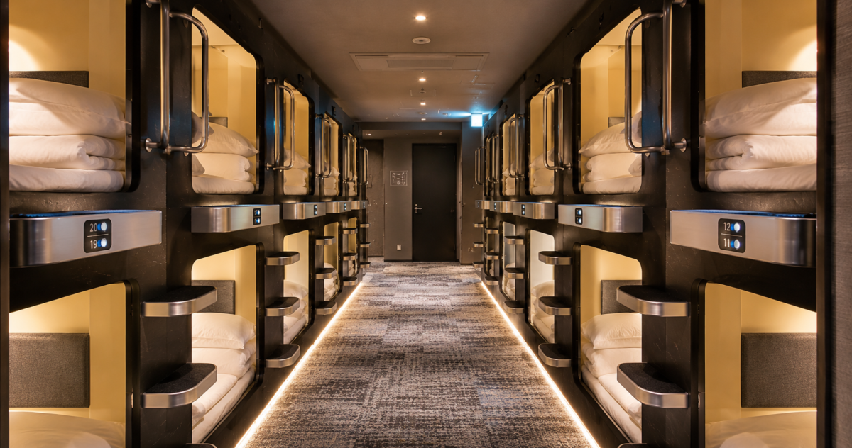 A modern Japanese capsule hotel corridor with rows of compact sleeping pods and neatly arranged white bedding