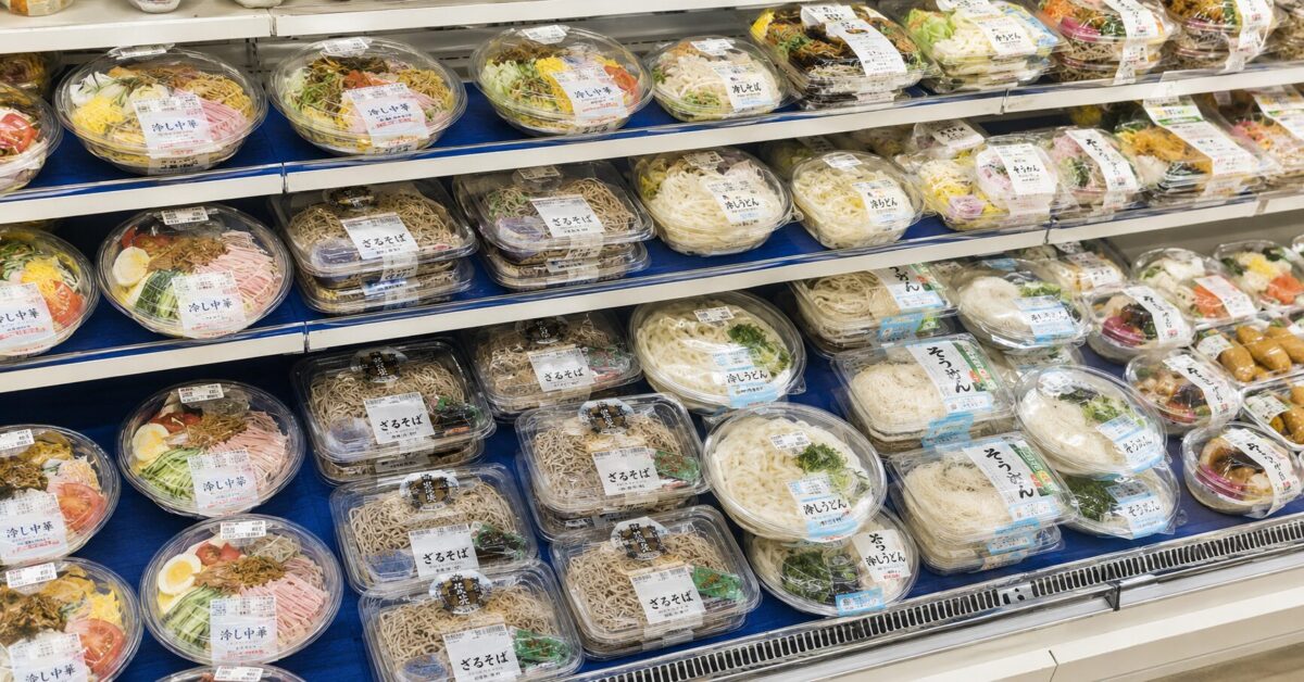 A refrigerated convenience store display filled with packaged hiyashi chuka, zaru soba, cold udon, and somen noodles.