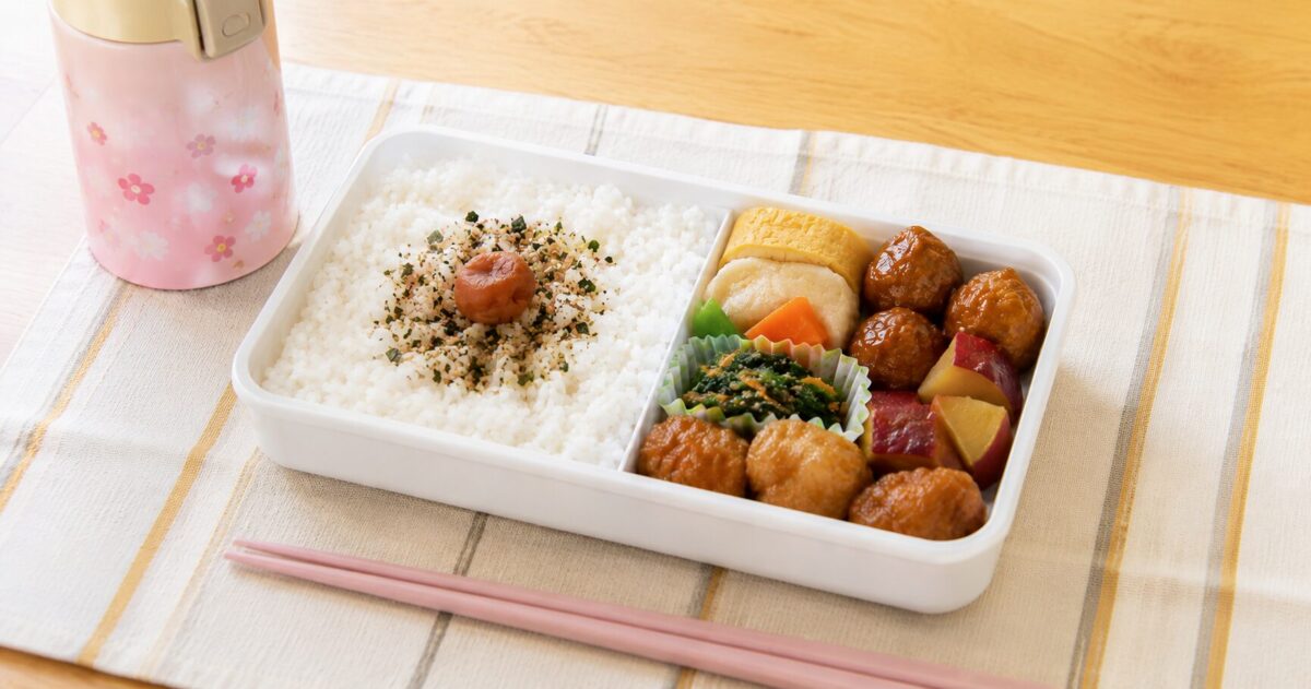 Homemade Japanese bento lunch with rice, meatballs, tamagoyaki, vegetables, chopsticks, and a thermos on a table.