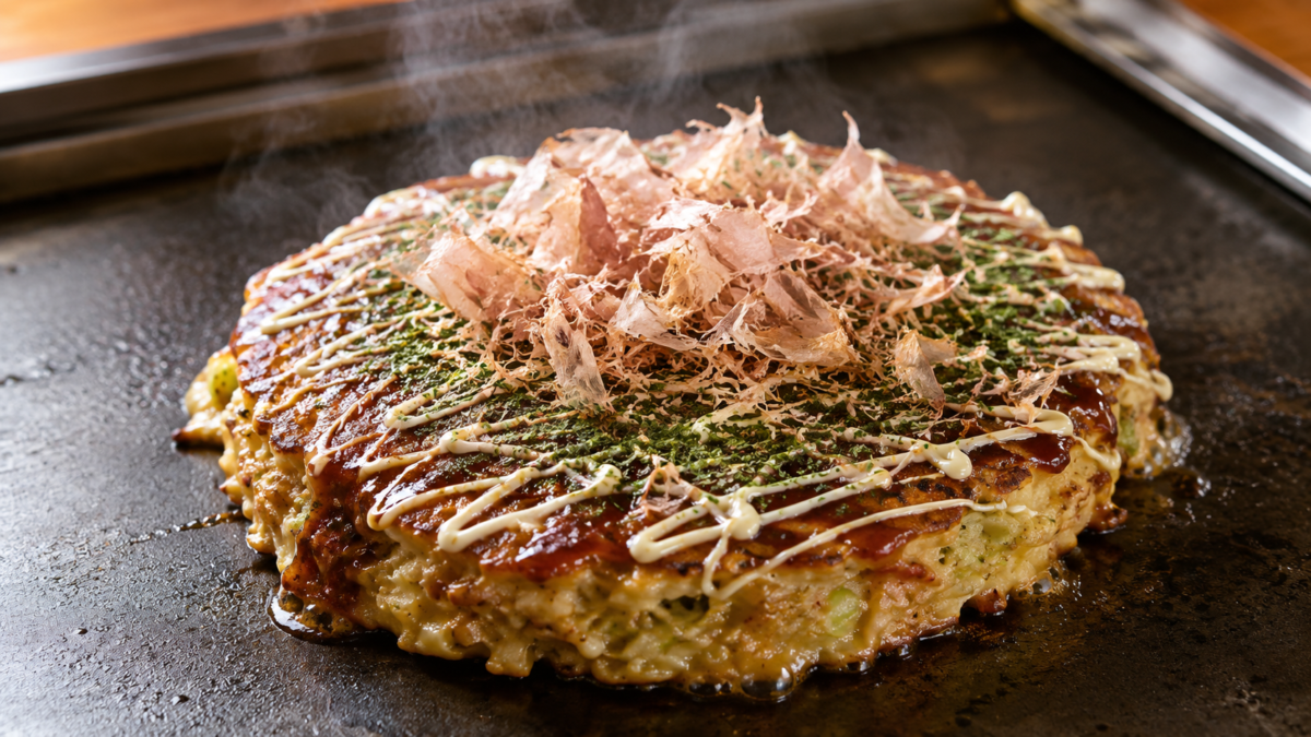 Freshly cooked Japanese okonomiyaki topped with sauce and bonito flakes on a hot plate