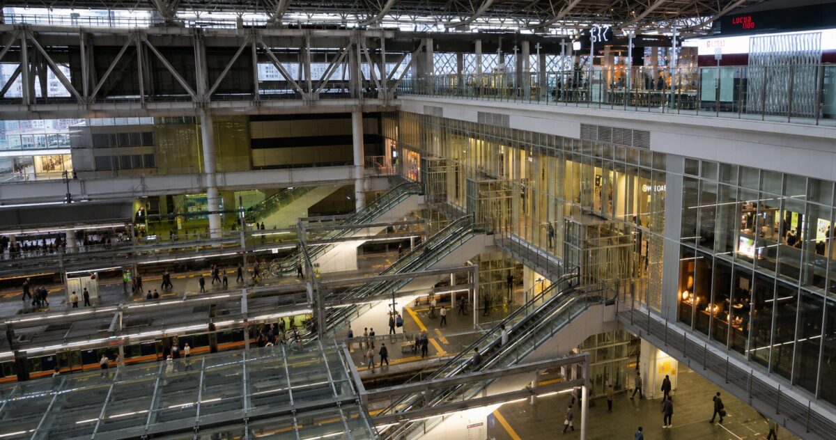 A panoramic view of Osaka Station showing railway platforms, the station concourse, escalators, and commuters in a modern transportation hub.