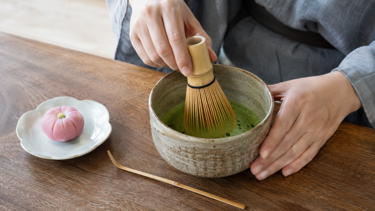 Matcha being whisked with a bamboo chasen in a traditional tea bowl beside a small Japanese sweet
